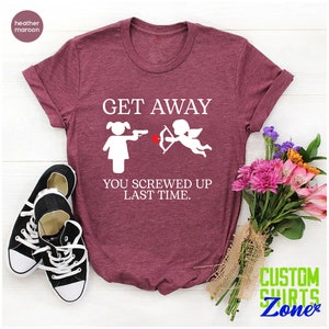 May include: A heather maroon t-shirt with the text "GET AWAY" in white. Below the text is a white illustration of a woman pointing a gun at a cupid with an arrow. The text "YOU SCREWED UP LAST TIME." is below the illustration.