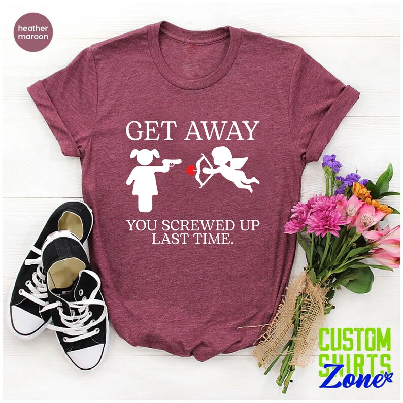May include: A heather maroon t-shirt with the text "GET AWAY" in white. Below the text is a white illustration of a woman pointing a gun at a cupid with an arrow. The text "YOU SCREWED UP LAST TIME." is below the illustration.