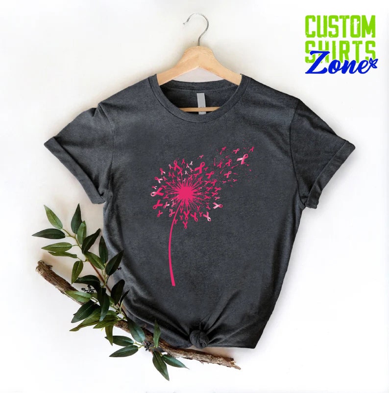 Op de afbeelding: Een donkergrijs T-shirt met een roze paardenbloemontwerp. De paardenbloem is gemaakt van roze linten, wat staat voor bewustwording van borstkanker. De tekst "CUSTOM SHIRTS Zone" staat in de rechterbovenhoek van de afbeelding.