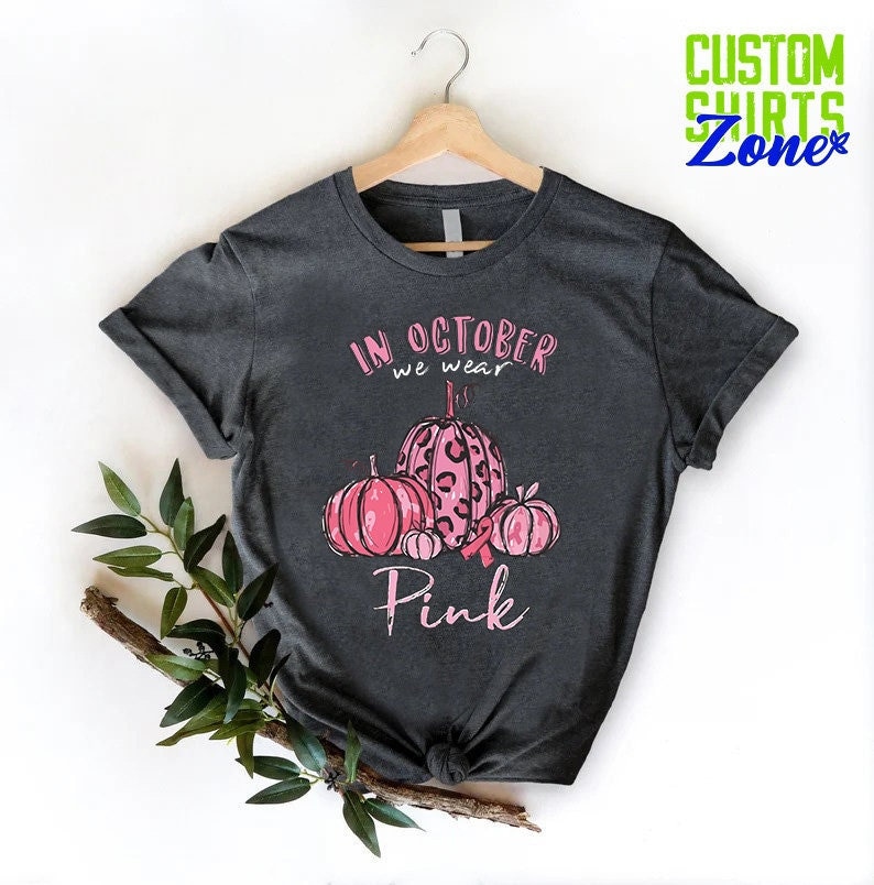 May include: A dark gray t-shirt with a pink and black pumpkin design. The text on the shirt reads "In October we wear Pink".