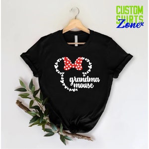 Op de afbeelding: Zwart T-shirt met een witte muizenkop, een rode strik met witte stippen en de tekst "grandma mouse". Het shirt hangt aan een houten hanger. De tekst "CUSTOM SHIRTS Zone" is zichtbaar in de rechterbovenhoek.