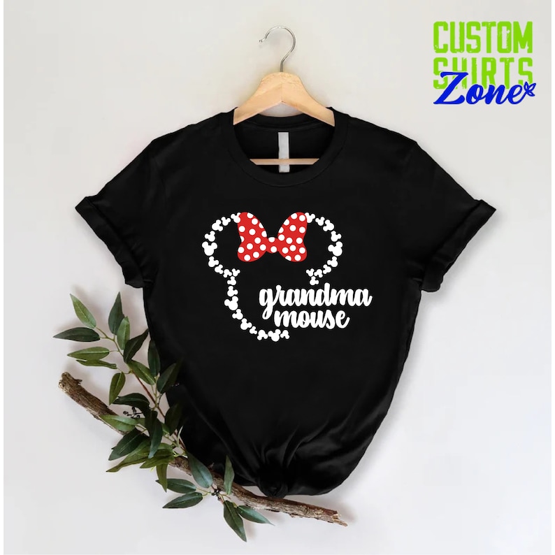 Op de afbeelding: Zwart T-shirt met een witte muizenkop, een rode strik met witte stippen en de tekst "grandma mouse". Het shirt hangt aan een houten hanger. De tekst "CUSTOM SHIRTS Zone" is zichtbaar in de rechterbovenhoek.