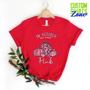 May include: Red t-shirt with a pink and black pumpkin design. The text on the shirt reads "In October we wear Pink".