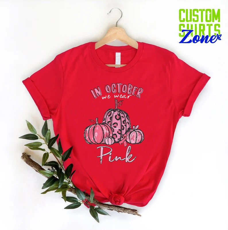 May include: Red t-shirt with a pink and black pumpkin design. The text on the shirt reads "In October we wear Pink".