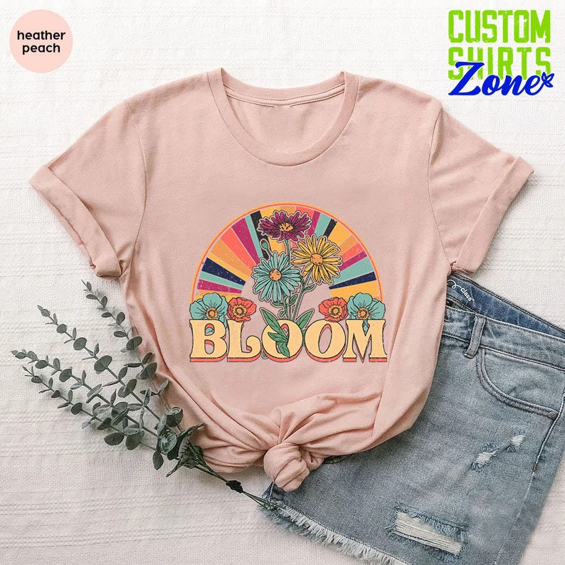 May include: A heather peach t-shirt with a retro-style graphic of flowers and a rainbow arch. The text "BLOOM" is printed in a vintage font below the graphic.