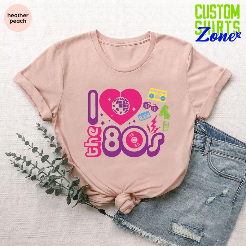 May include: A pink t-shirt with the text "I love the 80s" in a colorful retro font. The design includes a pink heart with a disco ball inside, a cassette tape, roller skates, sunglasses, and a record.