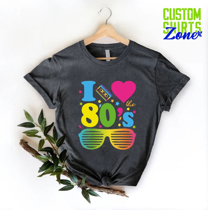 May include: A dark gray t-shirt with a graphic design that says "I love the 80's" with a pink heart, a cassette tape, and rainbow sunglasses.