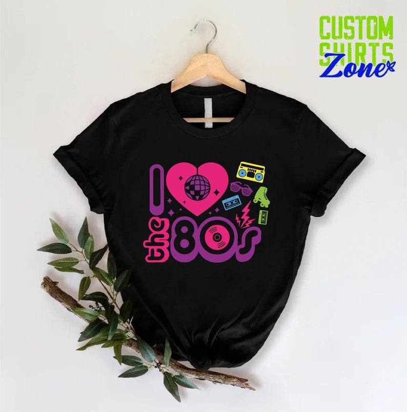 May include: Black t-shirt with a graphic design of a pink heart with a disco ball inside, the text "I love the 80s" in pink, purple, and blue, and 80s icons like roller skates, sunglasses, and a boombox.