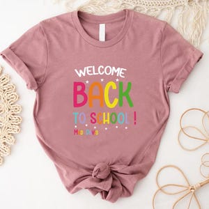 以下が含まれることがあります： 袖をまくったダスティローズ色のTシャツ。前面には、カラフルで遊び心のあるフォントで「WELCOME BACK TO SCHOOL！」の文字がプリントされています。テキストの下には「MS DVS」と書かれています。