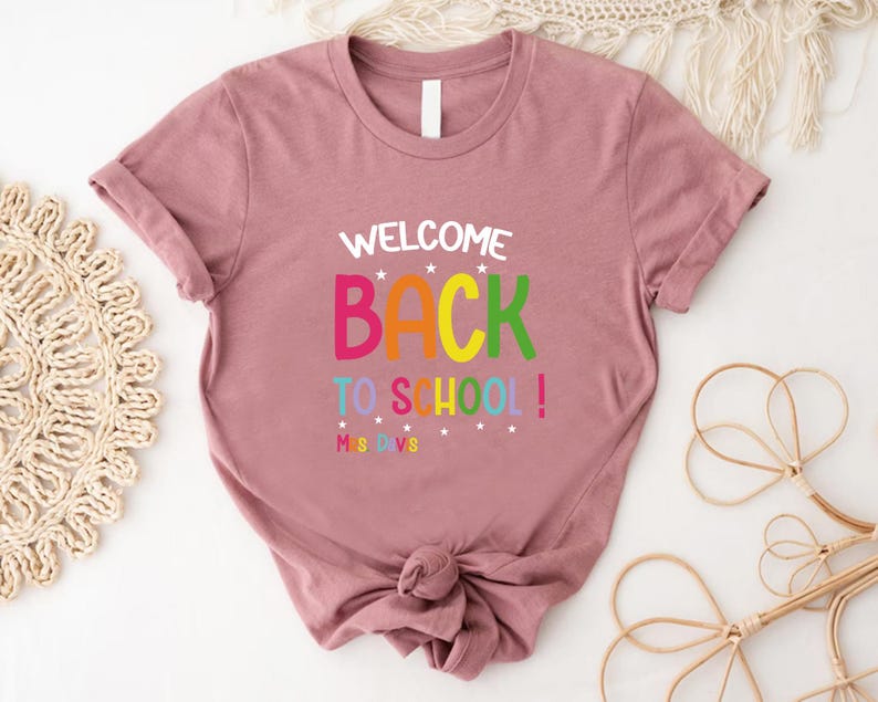 以下が含まれることがあります： 袖をまくったダスティローズ色のTシャツ。前面には、カラフルで遊び心のあるフォントで「WELCOME BACK TO SCHOOL！」の文字がプリントされています。テキストの下には「MS DVS」と書かれています。