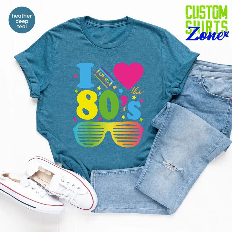 May include: A teal blue t-shirt with a graphic design that says "I love the 80's" with a pink heart, a cassette tape, and rainbow sunglasses.
