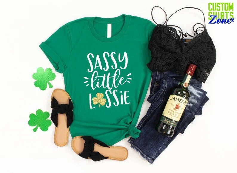 K&ouml;nnte beinhalten: Gr&uuml;nes T-Shirt mit dem Text "Sassy little Lassie" in Wei&szlig; mit einem goldenen Glitzer-Kleeblatt. Das Shirt wird mit einer blauen Jeans, einem schwarzen Spitzenoberteil, schwarzen Sandalen und einer Flasche Jameson Irish Whiskey kombiniert.