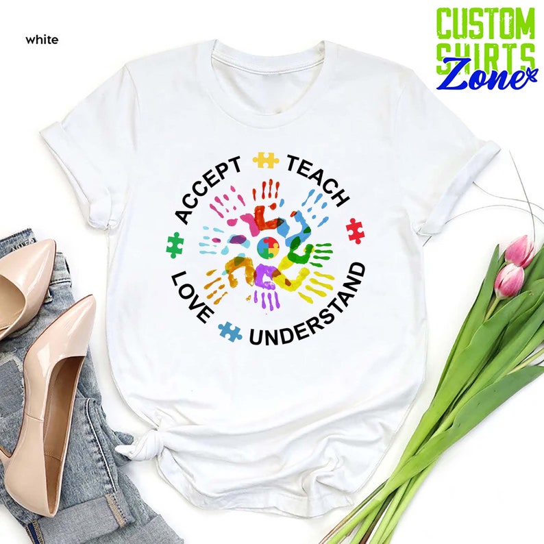 Puede incluir: Camiseta blanca con un dise&ntilde;o de huellas de manos de colores en el centro. Las huellas de manos est&aacute;n dispuestas en c&iacute;rculo con las palabras "Aceptar, Amar, Ense&ntilde;ar, Entender" que las rodean.