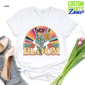 May include: White t-shirt with a retro-style graphic of a rainbow with flowers and the word "BLOOM" in yellow letters.
