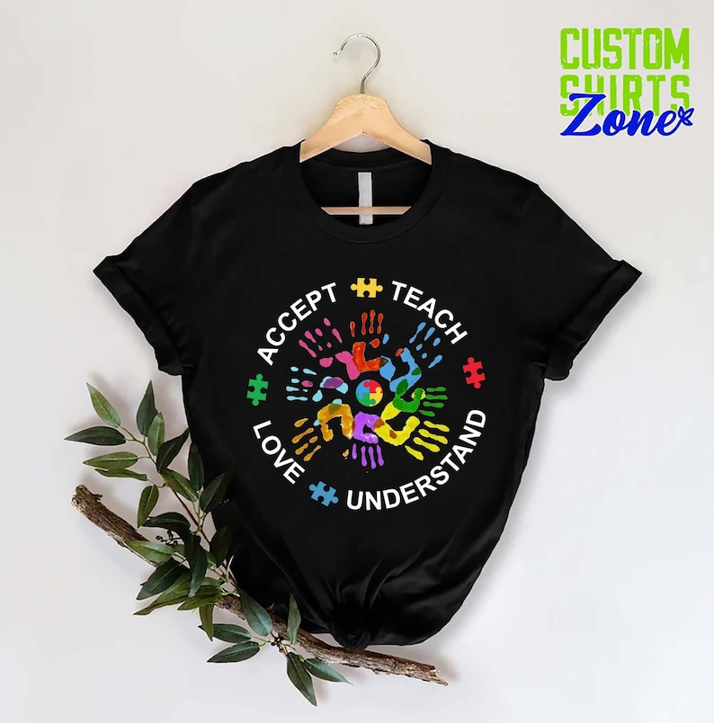 Puede incluir: Camiseta negra con un dise&ntilde;o circular blanco con las palabras "Aceptar, Ense&ntilde;ar, Amar, Entender" y un dise&ntilde;o de huellas de manos de colores con piezas de rompecabezas.