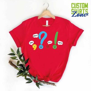 May include: Red t-shirt with a graphic of punctuation marks with faces and speech bubbles. The text in the speech bubbles reads "Wait", "What?", "Stop it", and "I agree!"