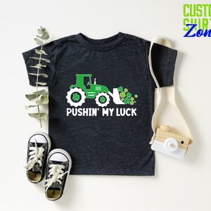 May include: A dark gray t-shirt with a green tractor and shamrocks. The text "Pushin' My Luck" is printed on the shirt.