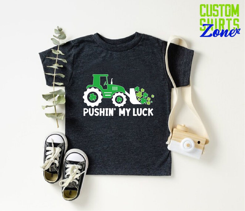 May include: A dark gray t-shirt with a green tractor and shamrocks. The text "Pushin' My Luck" is printed on the shirt.