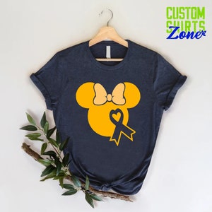 K&ouml;nnte beinhalten: Ein dunkelblaues T-Shirt mit einem gelben Mickey-Maus-Kopf-Design. Der Mickey-Maus-Kopf hat eine gelbe Schleife und ein blaues Band mit einem Herzen in der Mitte.