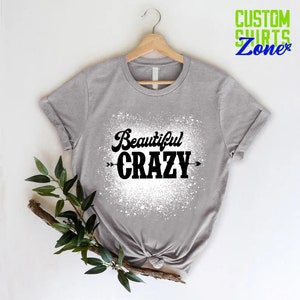 Puede incluir: Una camiseta gris con un dise&ntilde;o de salpicaduras blancas y el texto "Beautiful CRAZY" en negro con flechas a cada lado.