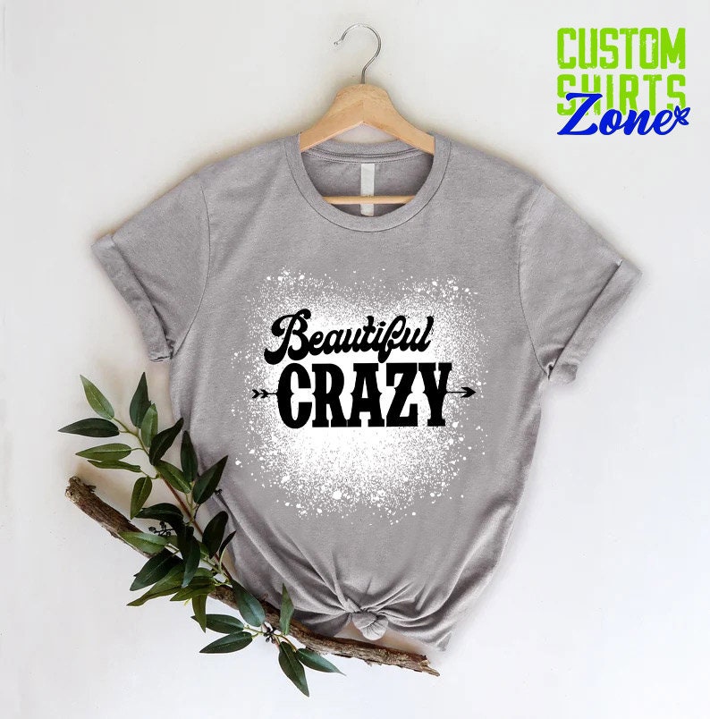 Puede incluir: Una camiseta gris con un dise&ntilde;o de salpicaduras blancas y el texto "Beautiful CRAZY" en negro con flechas a cada lado.