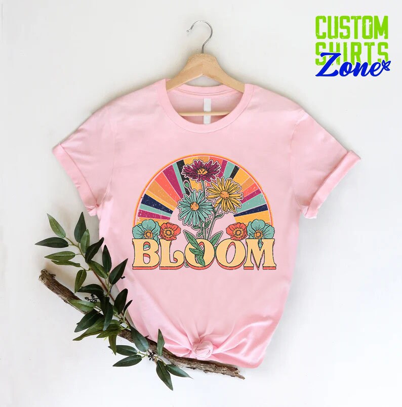 May include: Pink t-shirt with a retro-style graphic of flowers and a rainbow with the word "BLOOM" in a vintage font.