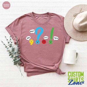 May include: A heather mauve t-shirt with a graphic of punctuation marks with faces. The punctuation marks are a comma, question mark, exclamation point, and period. The text on the shirt reads "What?", "Wait", "Stop it", and "I agree!"
