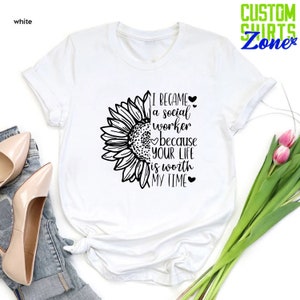 May include: White t-shirt with a black sunflower graphic and the text "I became a social worker because your life is worth my time".