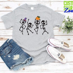May include: A gray t-shirt with a graphic of four skeletons dancing. The skeletons are wearing hats and one is holding a pumpkin. The text "CUSTOM SHIRTS Zone" is in the top right corner of the image.