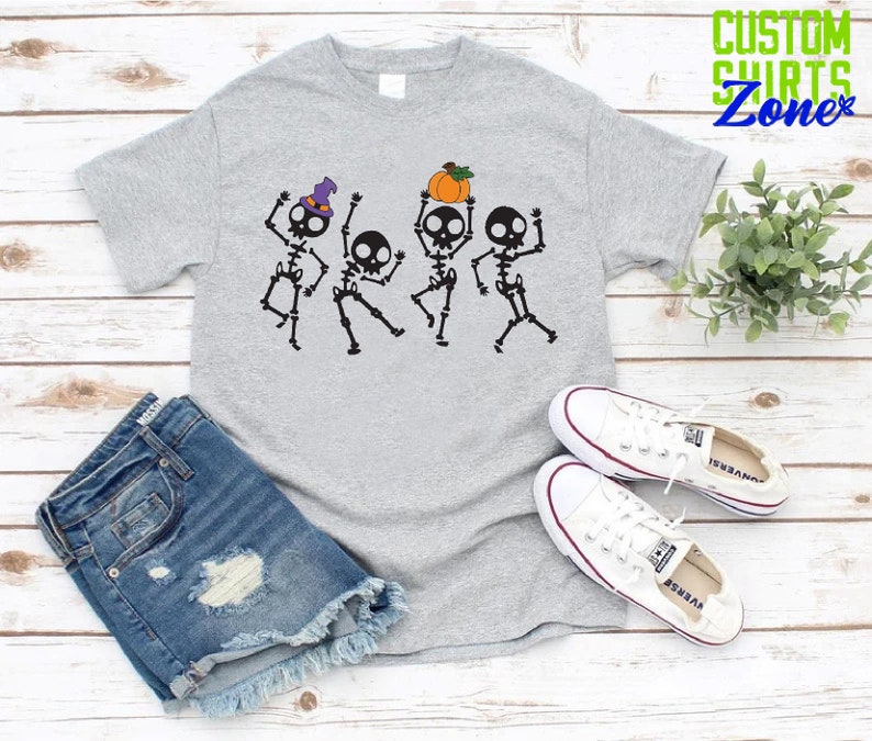 May include: A gray t-shirt with a graphic of four skeletons dancing. The skeletons are wearing hats and one is holding a pumpkin. The text "CUSTOM SHIRTS Zone" is in the top right corner of the image.