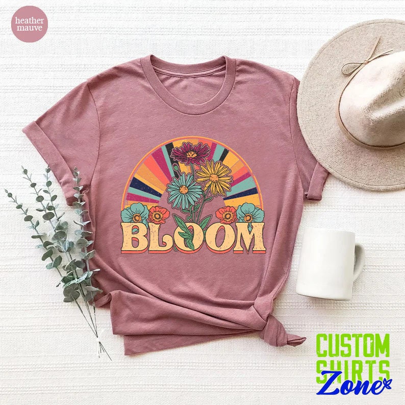 May include: A heather mauve t-shirt with a retro-style graphic of a bouquet of flowers with a rainbow arch and the word "BLOOM" in a vintage font.