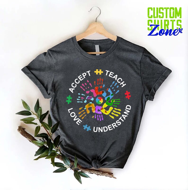 Puede incluir: Una camiseta gris oscuro con un dise&ntilde;o circular blanco con las palabras "Aceptar, Ense&ntilde;ar, Amar, Entender" y un patr&oacute;n de huellas de manos de colores. El dise&ntilde;o incluye piezas de rompecabezas.
