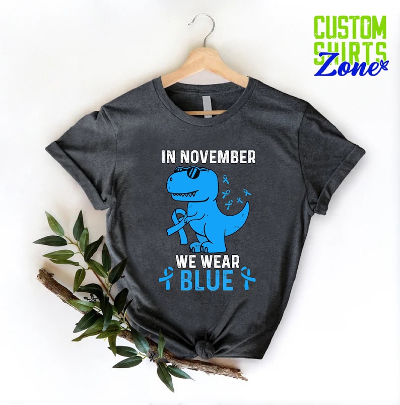 May include: Dark gray t-shirt with a blue dinosaur wearing sunglasses and holding a blue ribbon. The text "IN NOVEMBER WE WEAR BLUE" is printed above and below the dinosaur. The shirt is hanging on a wooden hanger.