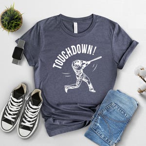 May include: A navy blue t-shirt with a white graphic of a baseball player swinging a bat and the text "TOUCHDOWN!" printed on the front.