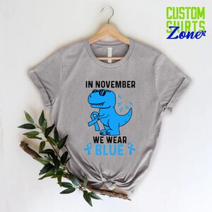 May include: A light gray t-shirt featuring a blue dinosaur wearing sunglasses and holding a blue ribbon. The text "IN NOVEMBER WE WEAR BLUE" is printed on the shirt, along with blue ribbons. The shirt is hanging on a wooden hanger.