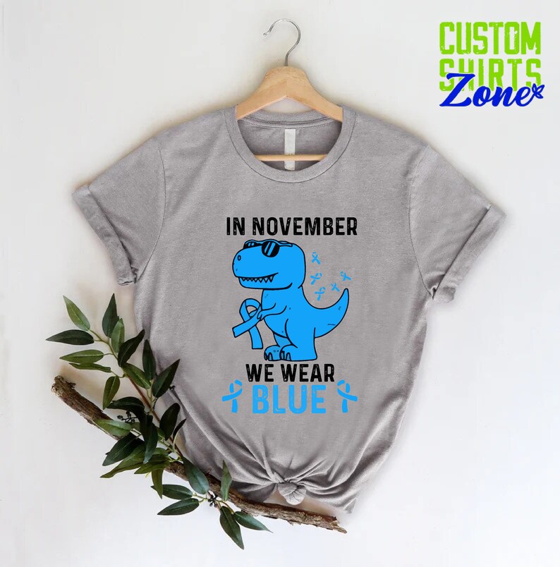 May include: A light gray t-shirt featuring a blue dinosaur wearing sunglasses and holding a blue ribbon. The text "IN NOVEMBER WE WEAR BLUE" is printed on the shirt, along with blue ribbons. The shirt is hanging on a wooden hanger.