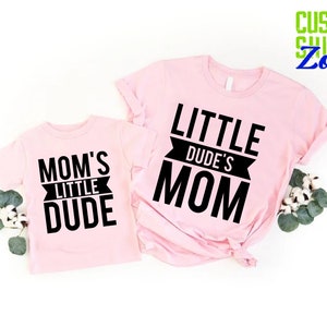 Op de afbeelding: Twee lichtroze t-shirts met zwarte tekst. E&eacute;n shirt zegt "MOM'S LITTLE DUDE" en de andere "LITTLE DUDE'S MOM". De shirts worden gepresenteerd op een witte ondergrond met groen.
