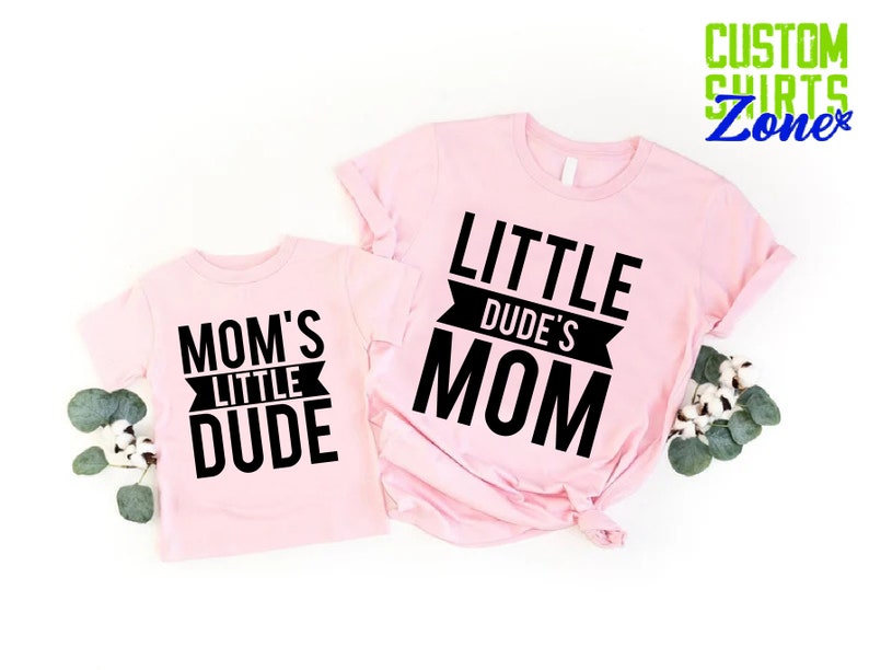Op de afbeelding: Twee lichtroze t-shirts met zwarte tekst. E&eacute;n shirt zegt "MOM'S LITTLE DUDE" en de andere "LITTLE DUDE'S MOM". De shirts worden gepresenteerd op een witte ondergrond met groen.