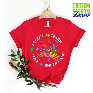 Puede incluir: Camiseta roja con un dise&ntilde;o circular blanco con las palabras "Aceptar", "Ense&ntilde;ar", "Amar" y "Entender" rodeadas de huellas de manos de colores y piezas de rompecabezas.