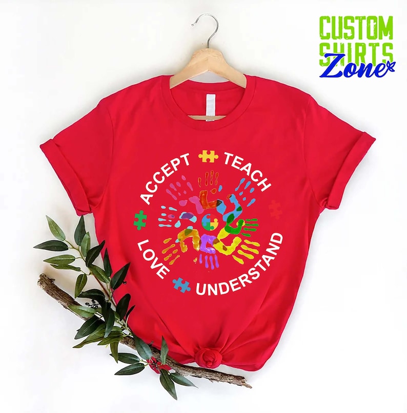 Puede incluir: Camiseta roja con un dise&ntilde;o circular blanco con las palabras "Aceptar", "Ense&ntilde;ar", "Amar" y "Entender" rodeadas de huellas de manos de colores y piezas de rompecabezas.