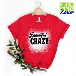 Puede incluir: Camiseta roja con un dise&ntilde;o de salpicaduras blancas y el texto "Beautiful CRAZY" en negro.