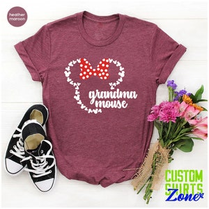 Op de afbeelding: Bordeauxrood t-shirt met een witte muizenkop en een rode stippenstrik, met de woorden "grandma mouse". Zwarte en witte sneakers staan linksonder, en een boeket bloemen staat rechts.
