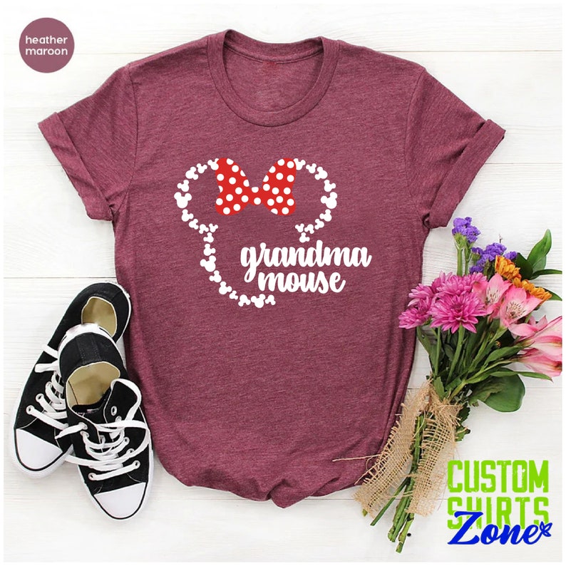 Op de afbeelding: Bordeauxrood t-shirt met een witte muizenkop en een rode stippenstrik, met de woorden "grandma mouse". Zwarte en witte sneakers staan linksonder, en een boeket bloemen staat rechts.