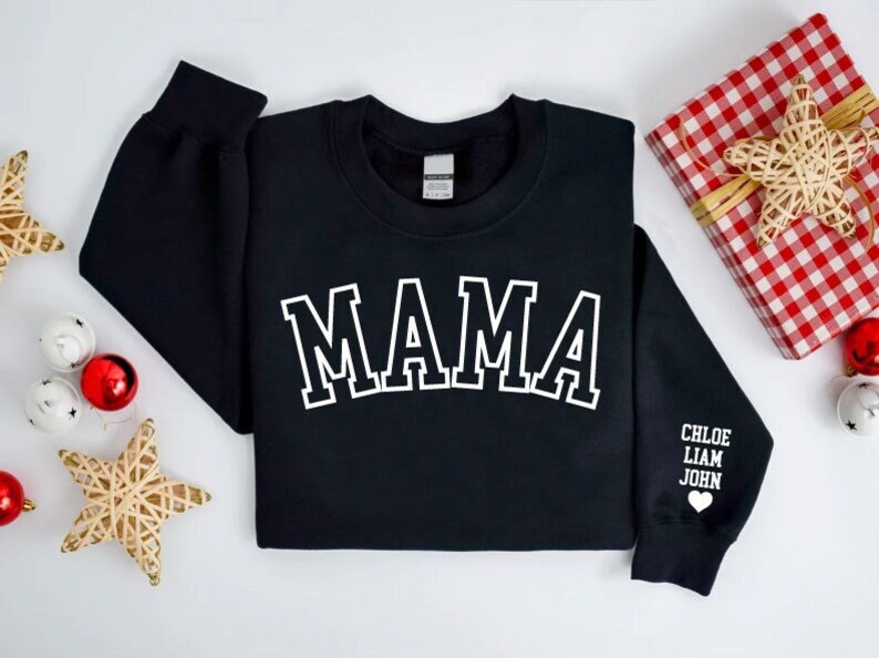 Puede incluir: Sudadera negra con la palabra "MAMA" en letras blancas con contorno en la parte delantera. La manga tiene los nombres "CHLOE, LIAM, JOHN" y un peque&ntilde;o coraz&oacute;n blanco.