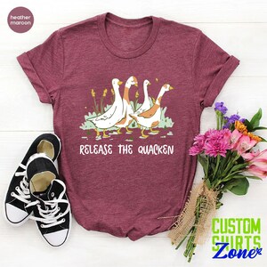 May include: A heather maroon t-shirt with a graphic of four geese walking in a line. The text "Release the Quacken" is printed below the geese.