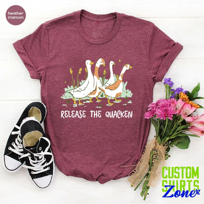 May include: A heather maroon t-shirt with a graphic of four geese walking in a line. The text "Release the Quacken" is printed below the geese.