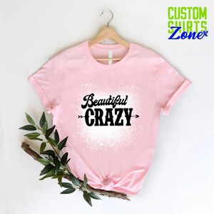 Puede incluir: Camiseta rosa con un dise&ntilde;o de salpicaduras blancas y el texto "Beautiful CRAZY" en negro con flechas a cada lado.