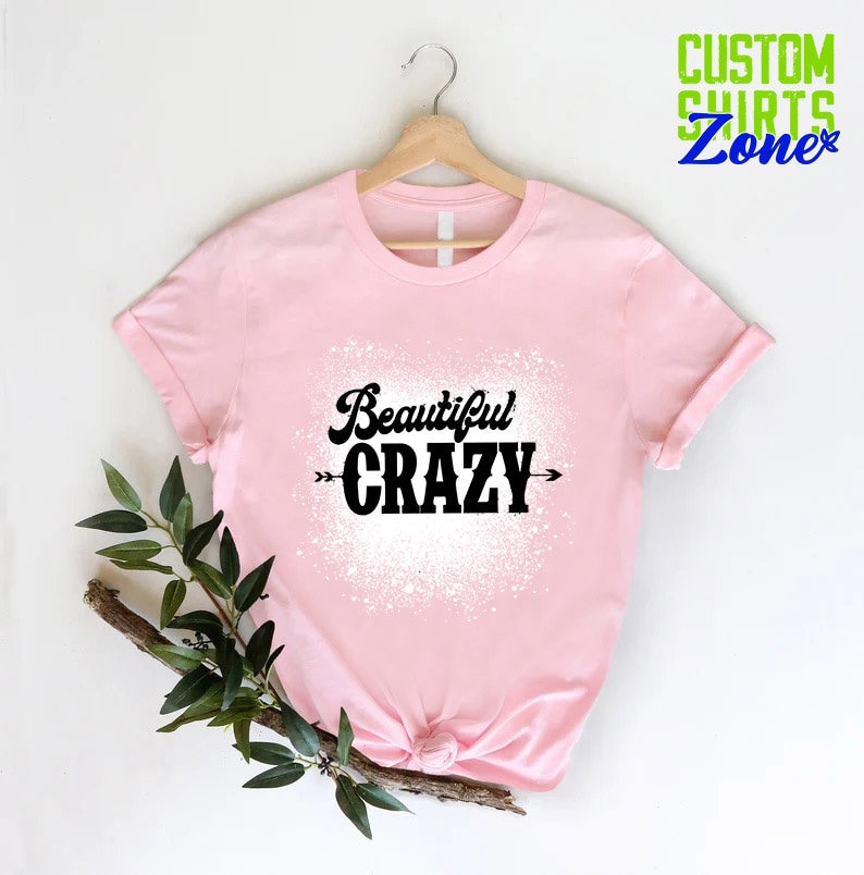 Puede incluir: Camiseta rosa con un dise&ntilde;o de salpicaduras blancas y el texto "Beautiful CRAZY" en negro con flechas a cada lado.