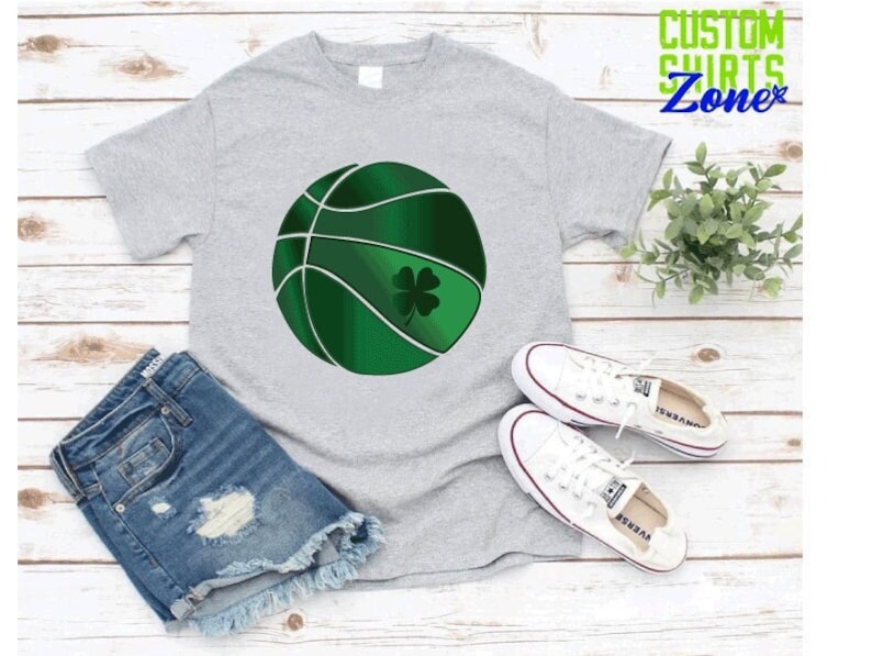 Puede incluir: Una camiseta gris con una pelota de baloncesto verde con un dise&ntilde;o de tr&eacute;bol de cuatro hojas. La camiseta est&aacute; sobre una superficie de madera blanca con una planta verde y zapatillas blancas.