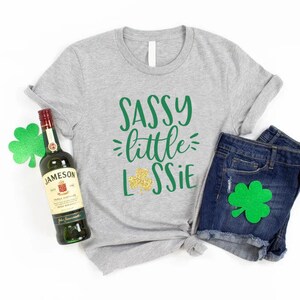 K&ouml;nnte beinhalten: Ein graues T-Shirt mit dem Text "Sassy little Lassie" in Gr&uuml;n und goldenen Glitzer. Das Shirt wird mit Jeans-Shorts und einer Flasche Jameson Irish Whiskey kombiniert. Es gibt gr&uuml;ne Kleebl&auml;tter, die um das Bild herum verstreut sind.